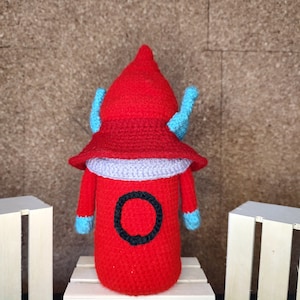 May include: A red crocheted plush toy shaped like a robot with blue arms and a purple hat. The toy has a black circle on its chest with a white outline.