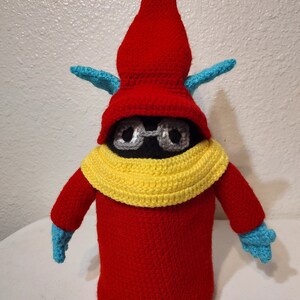 May include: A handmade crochet doll in the shape of a gnome. The doll has a red pointed hat, a black face with small silver glasses, and a yellow collar. The arms and hands are red with blue gloves.