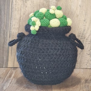 May include: A handmade crochet cauldron in black, filled with green and light green crochet balls. The cauldron has two black handles and is set against a wooden background. This whimsical piece is perfect for Halloween or fantasy-themed decor.