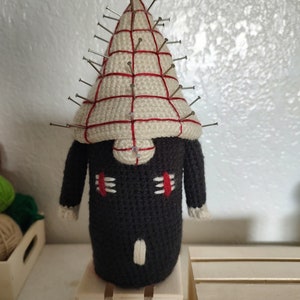 May include: A handmade crochet doll in the shape of a character with a white and red striped head, black body, and metal pins. The doll has small arms and a white and red face.
