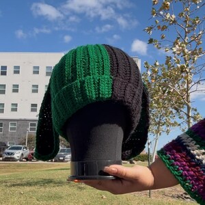 Puede incluir: Un gorro de ganchillo hecho a mano en verde y negro. El sombrero se exhibe en una cabeza de maniquí negra, sobre un fondo de cielo azul y un edificio. El gorro es un accesorio elegante.