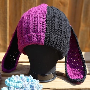 Pode incluir: Um chapéu de crochê feito à mão com um design dividido, com metade em roxo vibrante e a outra em preto. O chapéu apresenta longas orelhas de coelho flexíveis, perfeitas para um visual caprichoso. O chapéu é exibido em uma cabeça de manequim preta.