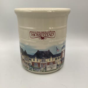 Pode incluir: Suporte para utensílios de cerâmica creme com um design Longaberger Homestead. O suporte apresenta uma cena pintada de um edifício com uma bandeira e as palavras "Longaberger Homestead" em uma faixa vermelha. O suporte tem formato cilíndrico.