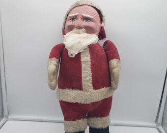 May include: A vintage Santa Claus doll with a red suit trimmed with white faux fur. The doll has a bald head with a white beard and blue eyes. Black boots complete the festive look, perfect for holiday decor.