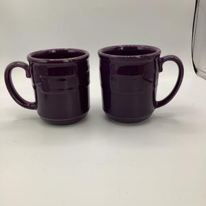 Puede incluir: Dos tazas de cerámica moradas a juego con un diseño texturizado y acanalado. Cada taza tiene un asa redondeada y una abertura ancha. Las tazas están sobre un fondo blanco liso.