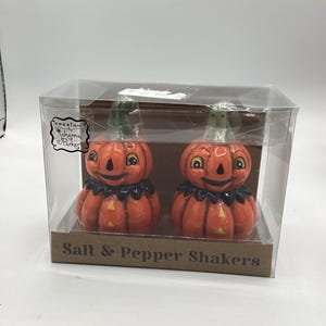 May include: A set of two orange ceramic pumpkin-shaped salt and pepper shakers with black accents, displayed in a clear plastic box. The shakers have smiling faces and green stems. The box has the text "Salt & Pepper Shakers".