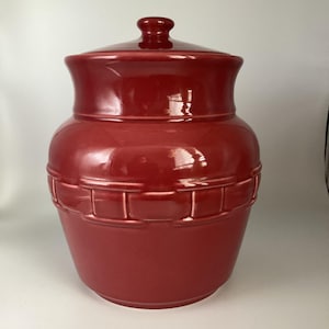 May include: A red ceramic cookie jar with a lid. The jar has a basket weave design around the middle and a rounded shape. The lid has a small knob on top.