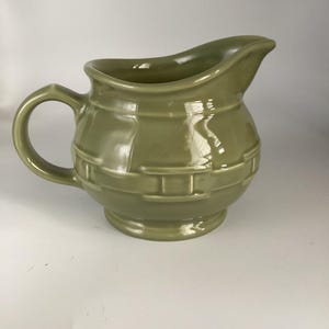 May include: A sage green ceramic pitcher with a curved spout and a rounded body. The pitcher has a handle and a decorative band around the middle. The pitcher is likely used for serving liquids.