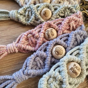 Könnte beinhalten: Fünf gehäkelte Schlüsselanhänger mit Holzperlen. Jede Perle hat ein Muschel-Design und den Text "Beste Mama". Die Schlüsselanhänger sind in verschiedenen Farben, darunter hellgrün, lila, rosa und weiß.