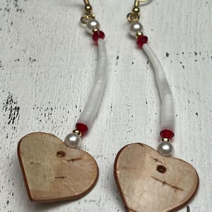May include: A pair of dangle earrings featuring heart-shaped wooden pendants. The earrings have gold-colored hooks, white curved beads, red beads, and small pearl beads. The wooden hearts have a natural wood grain.