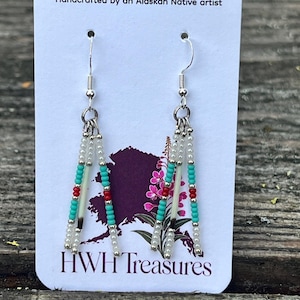 May include: Dangle earrings featuring three strands of beads in white, turquoise, and red, accented with silver-tone metal spacers and quill-like elements. The earrings are displayed on a white card labeled Handcrafted by an Alaskan Native artist and HWH Treasures with a purple graphic of the state of Alaska and a flower.