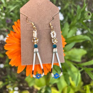 May include: A pair of dangle earrings featuring long white quill-like beads arranged in a triangular shape, accented with small cream, teal, and blue flower-shaped beads, gold-tone metal spacers, and gold-tone fishhook ear wires on a brown card.