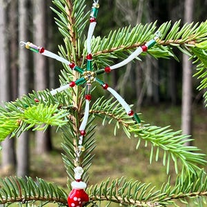 May include: A star-shaped ornament crafted from white, green, and red beads with a central gold-toned accent. A dangling charm shaped like a red mitten with white polka dots hangs from the bottom point, accented by clear and red beads.