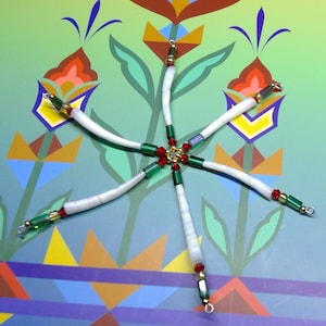 Può includere: Un ornamento a forma di stella fatto a mano con segmenti bianchi e curvi che si irradiano da un punto centrale. I segmenti sono adornati con perline rosse, verdi e dorate. L'ornamento è posto su uno sfondo colorato con motivi floreali.