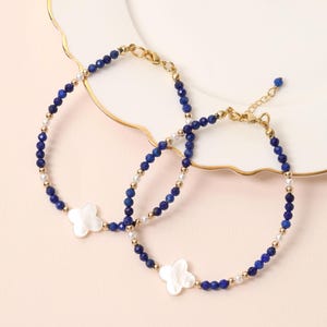May include: Two beaded bracelets with navy blue and gold beads, and white clover-shaped charms. The bracelets are on a white plate with a gold rim, set against a light pink background. The bracelets are delicate and elegant.