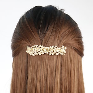 May include: A gold-colored hair barrette with a leaf design is set in brown hair. The barrette is rectangular and features a series of detailed leaves. The hair is straight and parted in the middle, with the barrette positioned horizontally.