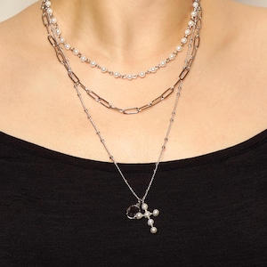 May include: A layered necklace set featuring three strands. The top two strands are delicate, with small pearls. The bottom strand has a chain with a cross pendant adorned with pearls. The jewelry is silver-toned and worn against a black top.