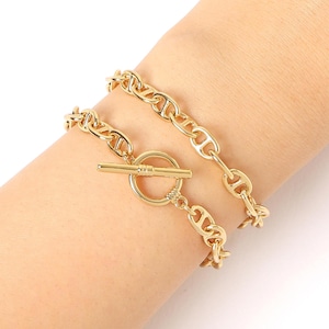 May include: Two gold chain bracelets with a toggle clasp. The bracelets are layered on a person's wrist.