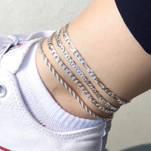 May include: A close-up of multiple silver anklets. The anklets feature different chain styles, including a rope chain and a flat curb chain. The anklets are worn on an ankle, with a white sneaker visible in the background.