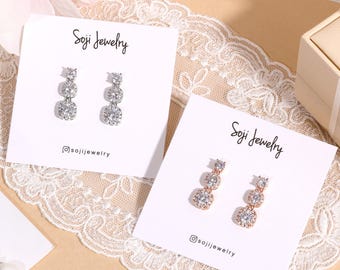 May include: Two pairs of Soji Jewelry earrings on white cards. One pair is silver-toned, the other is rose gold. Each earring features three graduated, square-cut, clear gemstones. The cards rest on a cream-colored lace surface.