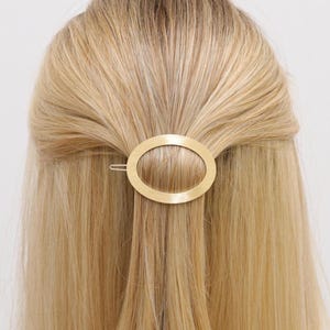 May include: A gold-colored oval hair clip is positioned in the hair. The hair is long, straight, and blonde. The clip is a simple, elegant accessory. The hair is styled in a half-up, half-down style.