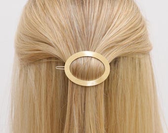May include: A gold-colored oval hair clip is positioned in the hair. The hair is long, straight, and blonde. The clip is a simple, elegant accessory. The hair is styled in a half-up, half-down style.