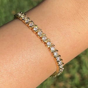 May include: A gold bracelet featuring a series of clear, round gemstones arranged in a wavy design. The bracelet is worn on a wrist, with a blurred green background.
