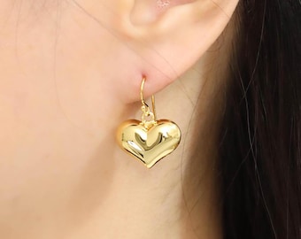 May include: A close-up of a gold heart-shaped earring. The earring is a shiny, solid gold color and has a rounded, puffy heart design. The earring is hanging from a gold hook.