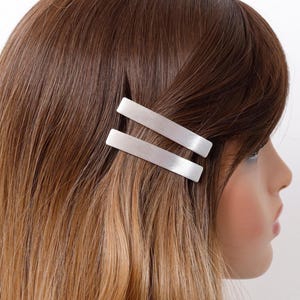 May include: Two silver barrettes are placed in brown and blonde hair. The hair is styled with a side part. The barrettes are rectangular and have a smooth, metallic finish. The background is white.