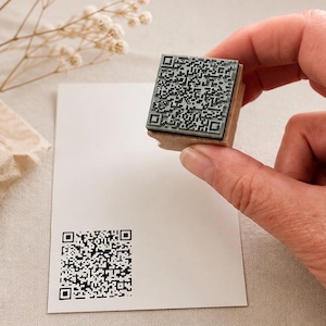 Könnte beinhalten: Ein QR-Code-Stempel mit Holzgriff wird über einer weißen Karte mit einem gestempelten QR-Code gehalten. Der Stempel ist quadratisch mit einem detaillierten schwarz-grauen QR-Code-Design. Die Karte ist leer und einsatzbereit.