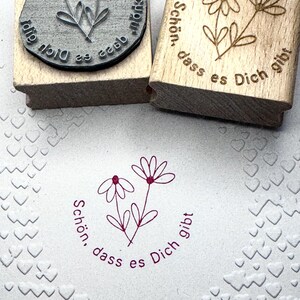 May include: Two wooden stamps with the text "Schon, dass es Dich gibt" and a simple flower design. The stamps are on a white surface with a heart pattern.