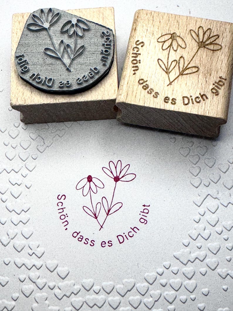 May include: Two wooden stamps with the text "Schon, dass es Dich gibt" and a simple flower design. The stamps are on a white surface with a heart pattern.