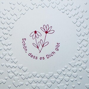 May include: A white card with a pink embossed circle of hearts. Inside the circle is a pink drawing of two flowers with the text "Sch&ouml;n, dass es Dich gibt".