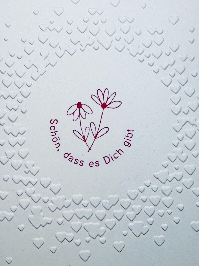 May include: A white card with a pink embossed circle of hearts. Inside the circle is a pink drawing of two flowers with the text "Sch&ouml;n, dass es Dich gibt".