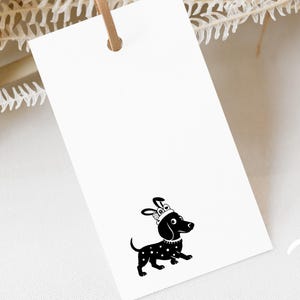 Könnte beinhalten: Ein weißes rechteckiges Etikett mit einem braunen Lederband. Das Etikett zeigt eine schwarz-weiße Illustration eines Dackels mit Krone und Tupfen. Der Hund geht.