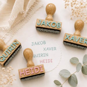 Könnte beinhalten: Holzstempel mit Namen in Hellblau, Rosa und Hellbraun. Die Stempel schreiben die Namen Jakob, Xaver, Quirin und Heidi. Die Namen sind auch auf einem weißen Papierkreis aufgedruckt.
