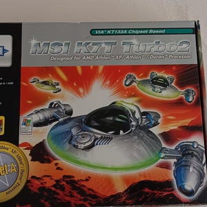 May include: A rectangular cardboard box for an MSI K7T Turbo2 motherboard featuring a graphic of a silver, saucer-shaped spacecraft against a red landscape. The box displays text including MSI K7T Turbo2, VIA KT133A Chipset Based, and Socket A.