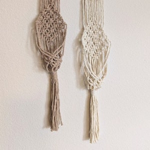 May include: Two macrame plant hangers with wooden rings at the top. The hanger on the left is brown and the hanger on the right is white.