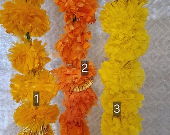 May include: Three strands of artificial marigold garlands in shades of yellow and orange. The garlands are made of layered, ruffled material, with some strands featuring gold accents. The garlands are likely used for decoration or religious ceremonies.
