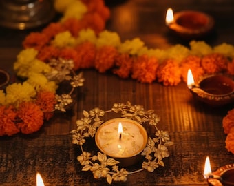 May include: A round gold-toned metal candle holder featuring a detailed floral wreath design surrounding a white wax candle with gold leaf accents, positioned among several lit clay oil lamps and garlands of orange and yellow marigold flowers.