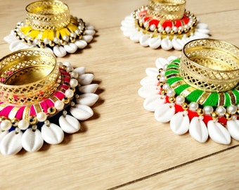 May include: Four decorative candle holders with gold-colored metal rims. Each holder features a colorful fabric base, adorned with white seashells and faux pearls. The bases are in yellow, red, pink, and green.
