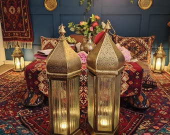 May include: Two tall, hexagonal gold-toned metal lanterns with intricate perforated patterns on the top sections and clear, vertically ribbed glass panels. Each lantern features a pointed roof and contains a small, glowing light inside.