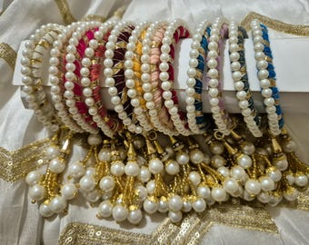 May include: A set of colorful beaded bangles with pearl accents and gold tassels. The bangles are made of thread and are decorated with stripes of different colors. The tassels are made of gold thread and are adorned with pearls.