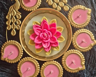 May include: A gold-toned metal circular tray featuring two peacock figures and a central pink and yellow lotus-shaped candle, surrounded by seven smaller round gold-toned candle holders containing pink wax candles with visible wicks.