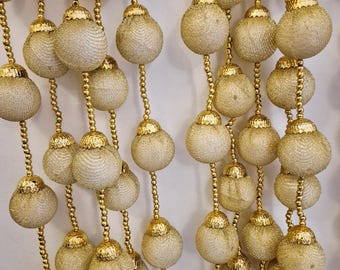 May include: Close-up of decorative gold beaded garland. The garland features multiple strands of small, round, textured, cream-colored orbs with gold caps, strung together with gold beads. The orbs are evenly spaced and hang vertically.