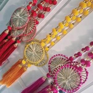 Puede incluir: Tres adornos colgantes decorativos con largas hileras de pompones en rojo, amarillo y magenta. Cada pieza incluye detalles circulares de metal tejido y largas borlas del mismo color en la parte inferior, creando un aspecto vibrante y superpuesto.