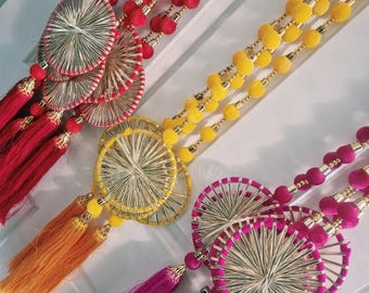 May include: Three decorative wall hangings featuring long strands of pom-poms in red, yellow, and magenta. Each piece includes circular woven metallic accents and long, matching colored tassels at the bottom, creating a vibrant, layered appearance.