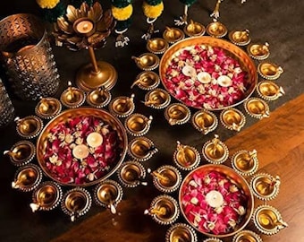 May include: Three round gold-toned metal bowls filled with pink and white flower petals, each surrounded by a ring of small attached oil lamp holders. A tall lotus-shaped candle holder and a metal lantern are positioned nearby.