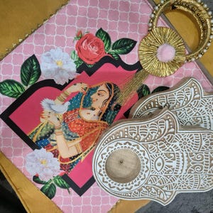 Puede incluir: Una bolsa de tela rosa y dorada con un diseño floral y un par de porta inciensos de madera blanca en forma de mano.