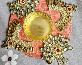 May include: A gold decorative tray with three ornate bird-shaped embellishments. Each bird is adorned with clear rhinestones and a red jewel in the center. The tray is set on a pink fabric background.